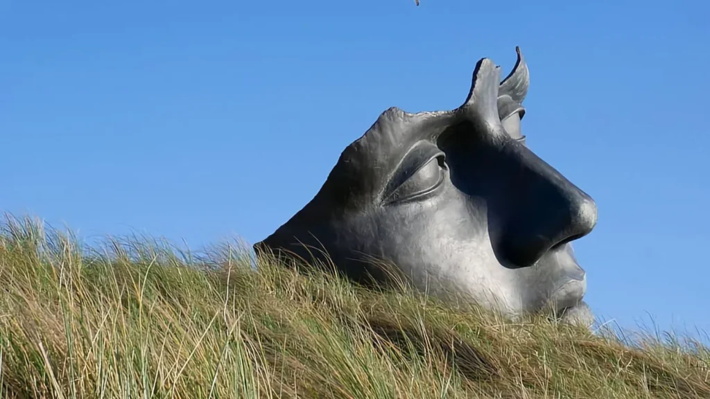 A large bronze sculpture of a face lying among dune grass against a blue sky