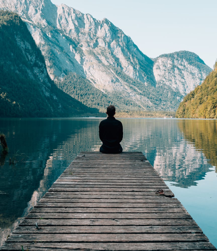 A solitary figure sits in meditation at the end of a wooden pier extending into a calm alpine lake, surrounded by towering mountains and lush forests. The serene scene captures the essence of mindfulness and nature's tranquility.