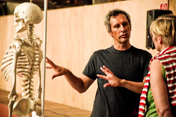 Malcolm Manning at ImPulsTanz Vienna, explaining a movement with open hands beside an anatomical skeleton, talking with a workshop participant