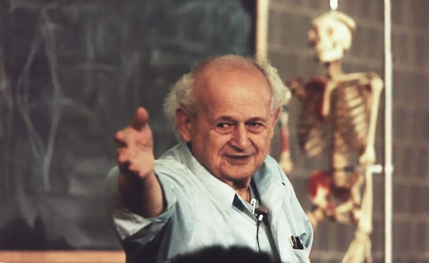 Moshe Feldenkrais teaching a training group, gesturing towards the class, with an anatomical skeleton behind him